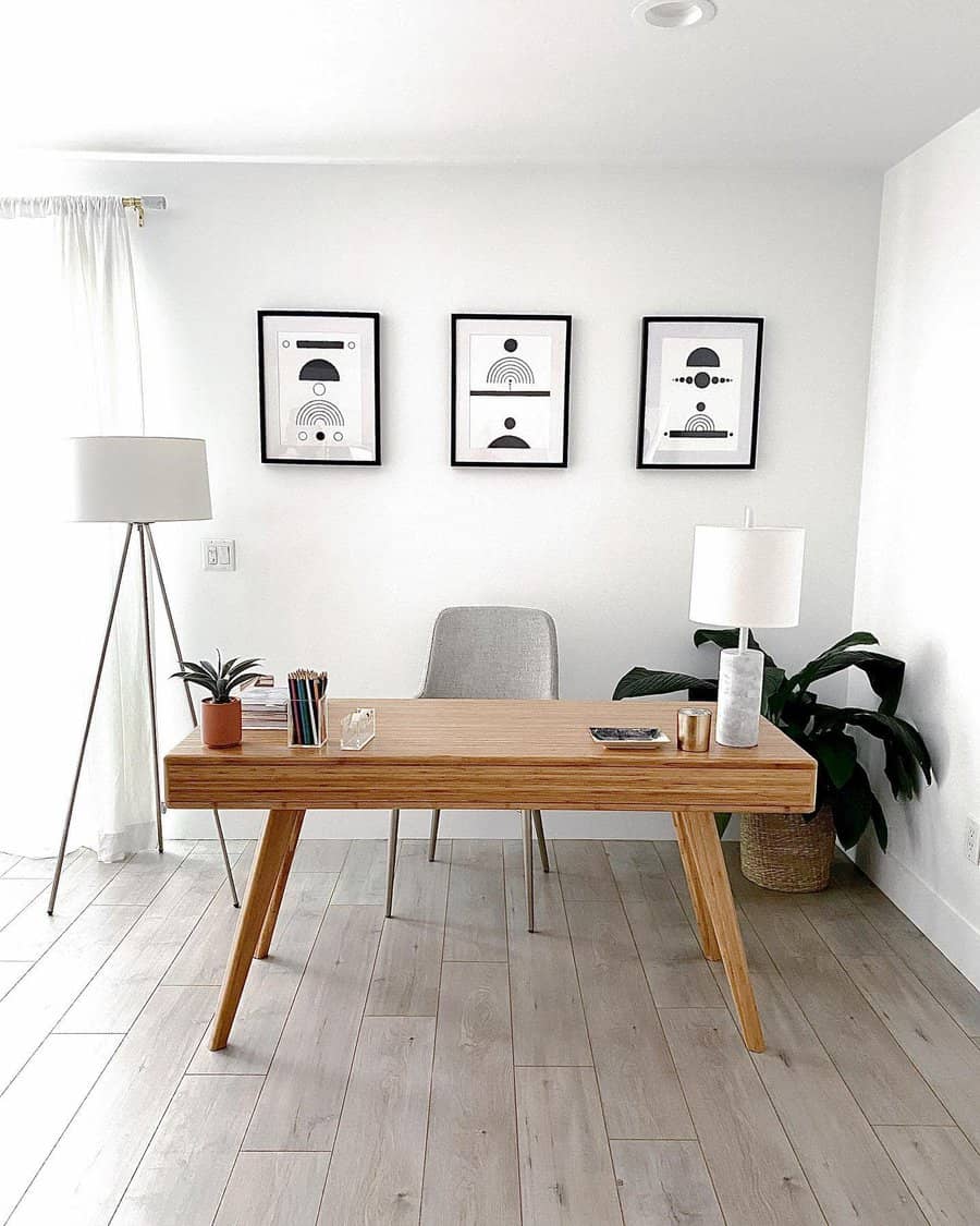 Minimalist home office with a wooden desk, chair, floor lamp, plant, and three abstract art pieces on a white wall