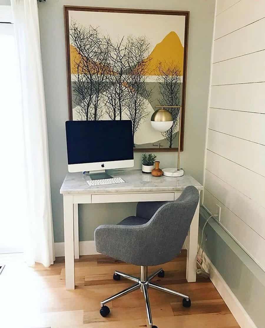 Compact home office with a computer on a white desk, gray chair, and art piece of trees and mountains in orange tones on the wall