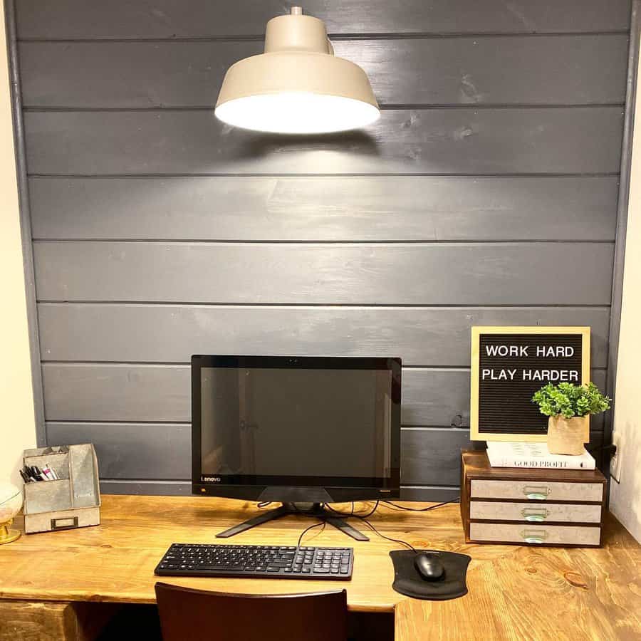 Minimalist desk setup with a computer, keyboard, lamp, and plant featuring a sign that says "WORK HARD PLAY HARDER" on a wood desk against a gray paneled wall