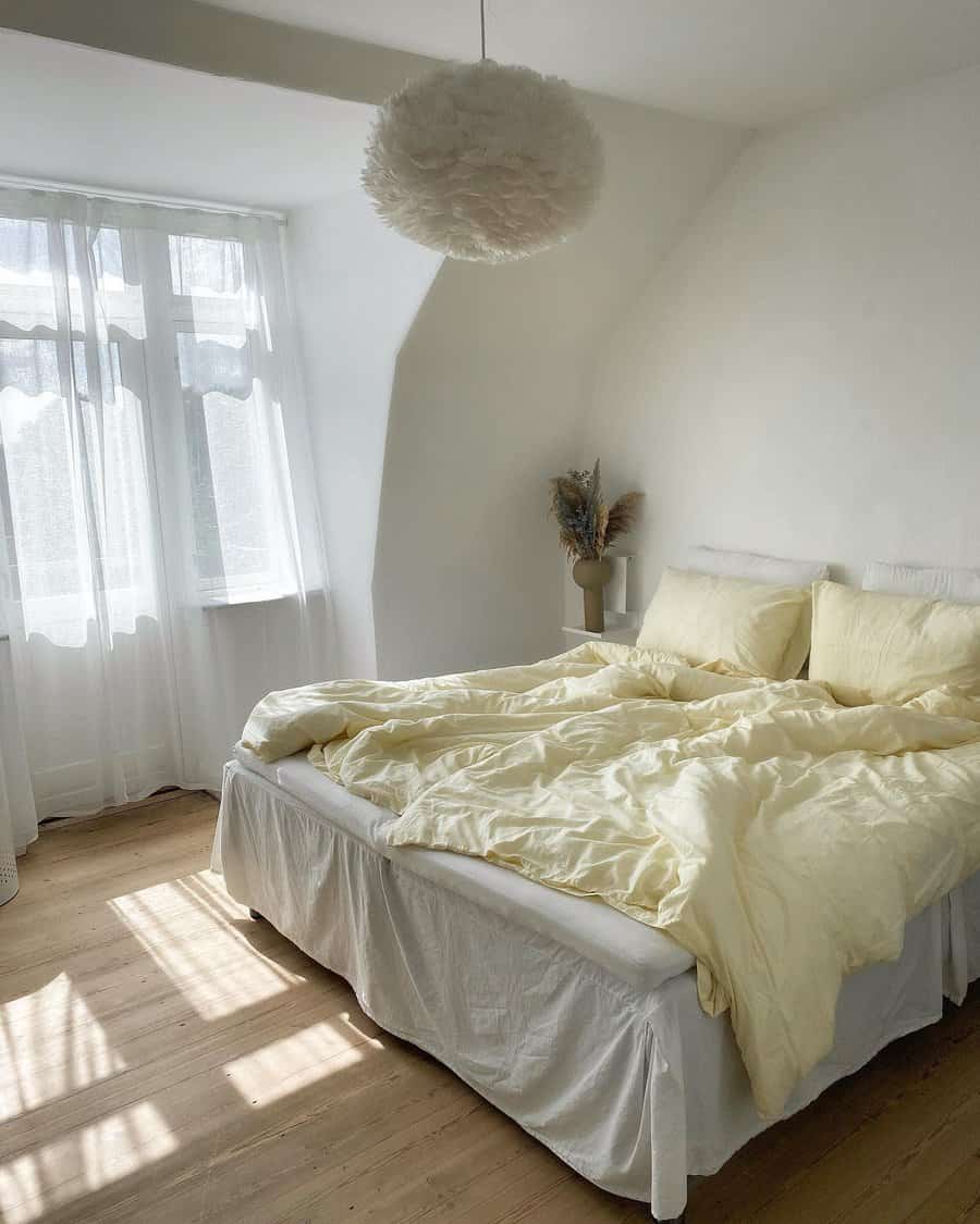 Bright bedroom with white walls, soft yellow bedding, sheer curtains, and a fluffy ceiling light; sunlight streams through the window