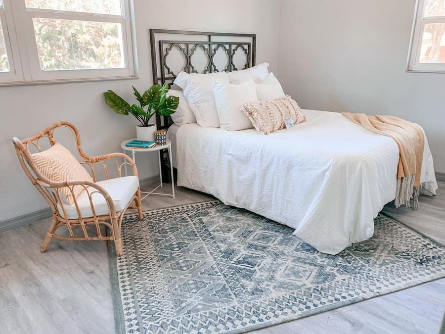 A cozy bedroom with a white bed, decorative pillows, a tan throw, a wicker chair, a plant, and a patterned rug on wooden floors