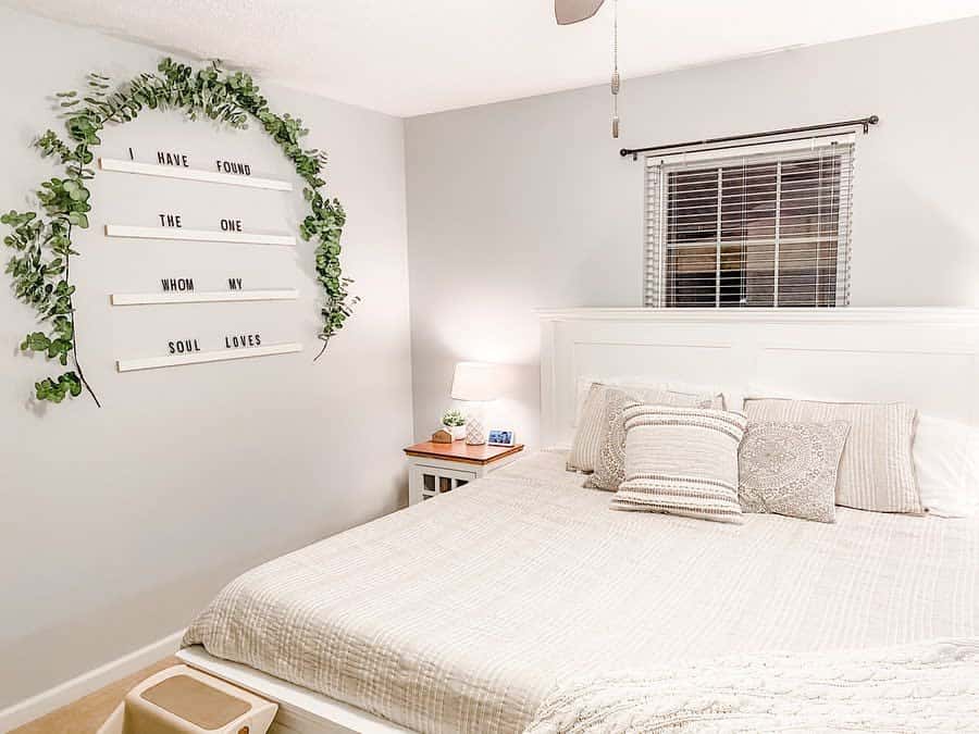 Bedroom with a white bed, decorative pillows, a nightstand with a lamp, and a wall sign with greenery; cozy and minimalist decor