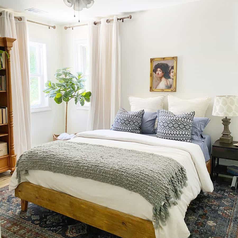 Cozy bedroom with a wooden bed, gray blanket, decorative pillows, a potted plant, and artwork on the wall