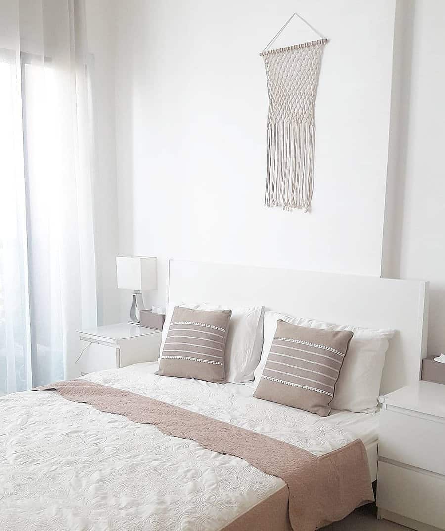 Minimalist bedroom with white bed, beige pillows, macramé wall hanging, and soft lighting from a window with sheer curtains