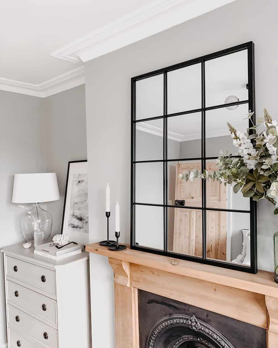 Living room with a modern mirror, white dresser, lamp, candles, and a wooden fireplace mantel; decor includes plants and a framed picture