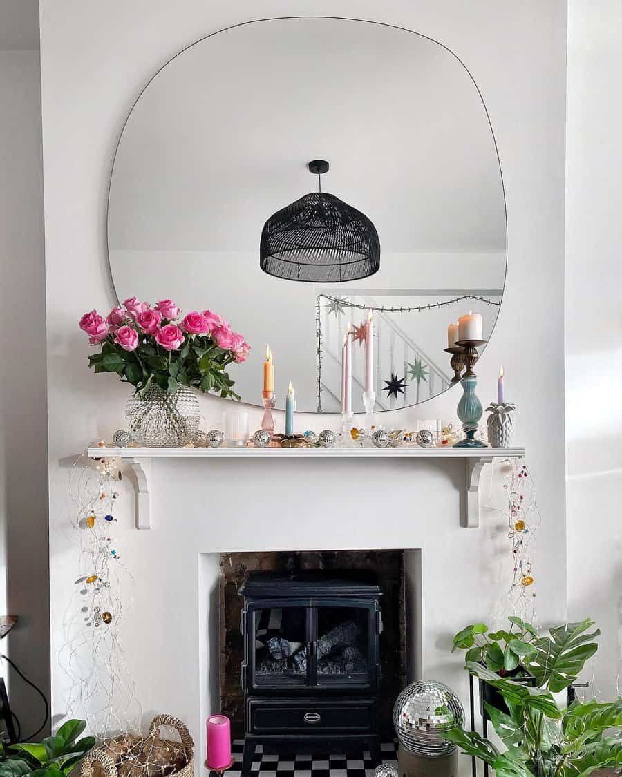 A fireplace with a large, irregular mirror above, featuring decor that includes pink roses, candles, string lights, and greenery in a cozy setup