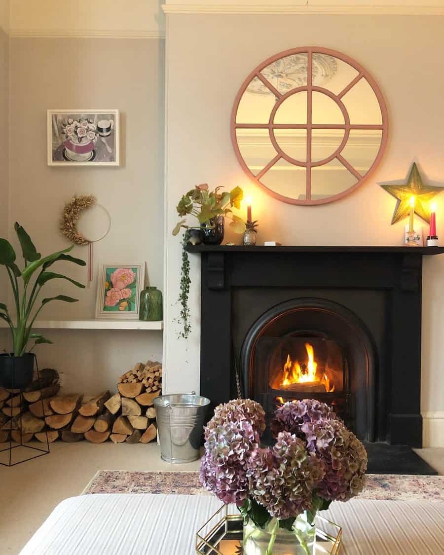 Cozy living room with a lit fireplace, circular mirror above, potted plants, stacked firewood, and a vase of hydrangeas on a table