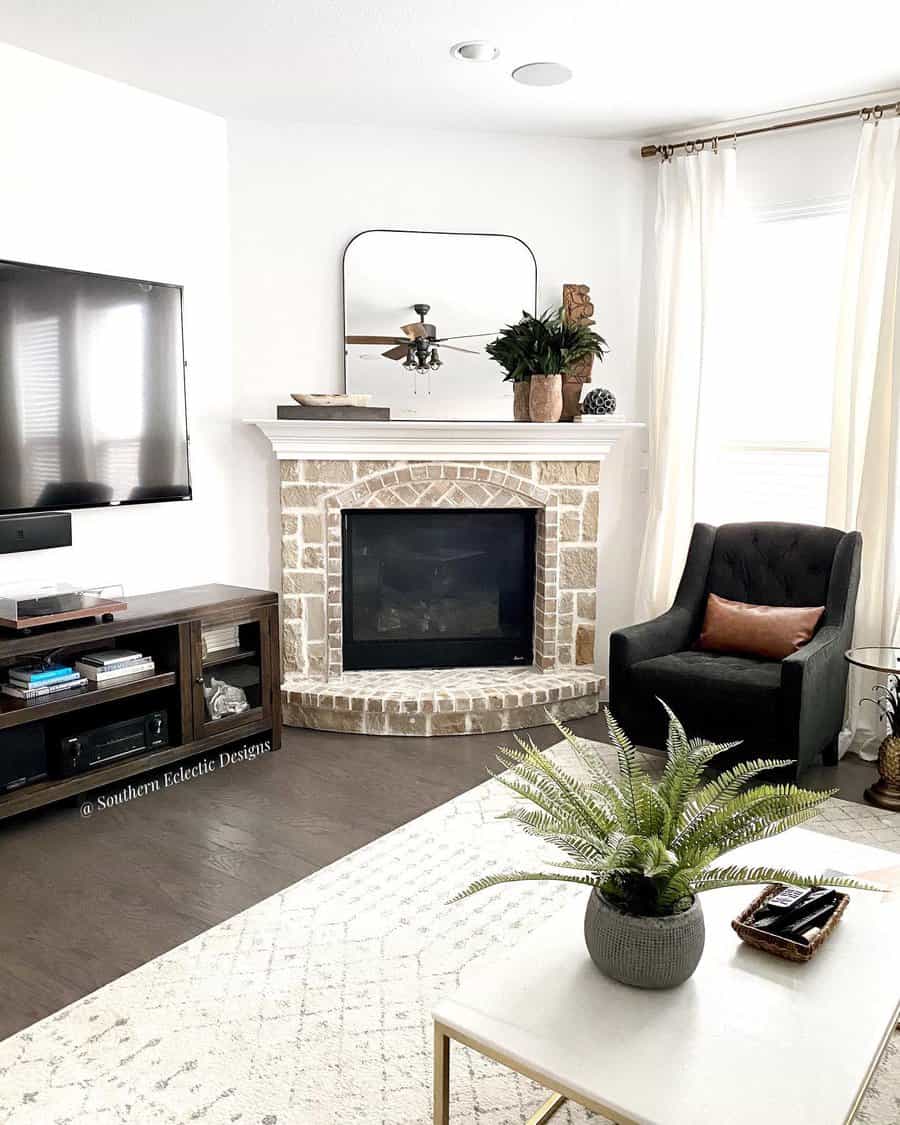 Living room with a brick fireplace, mounted TV, armchair, potted fern on coffee table, mirror above mantle, and white curtains