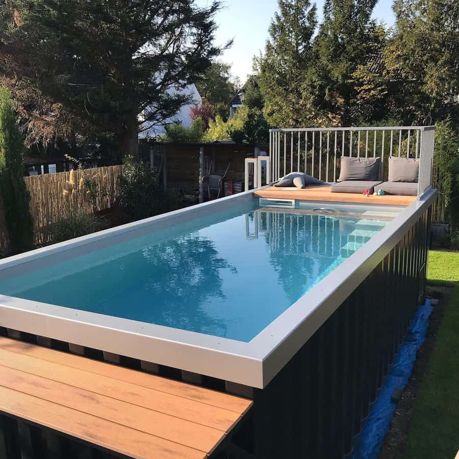 A rectangular above-ground pool with a deck and seating, surrounded by trees and grass, it reflects the blue sky above