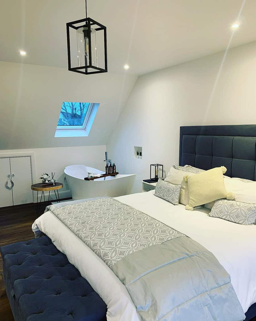 Luxurious attic bedroom with a deep blue tufted bed, freestanding bathtub, and skylight. Modern lighting and soft textures create a spa-like retreat.