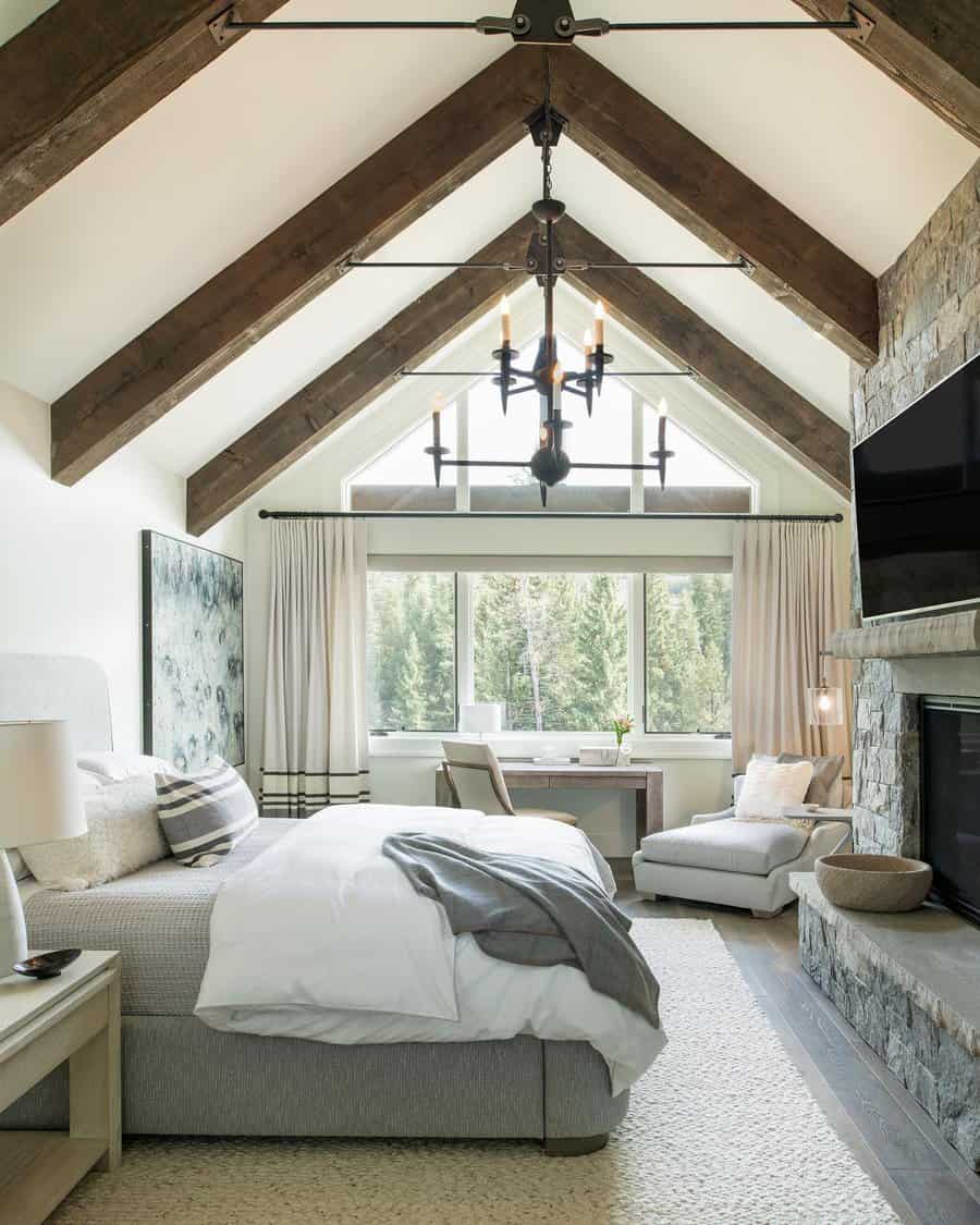 Elegant attic bedroom with exposed wood beams, a stone fireplace, and floor-to-ceiling windows. Neutral tones and soft textures create a cozy retreat.