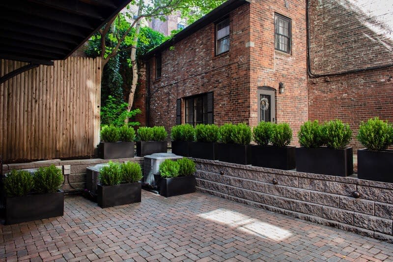 Stylish urban patio with interlocking brick pavers, modern black planters, lush greenery, and a rustic brick facade for a cozy and contemporary outdoor space