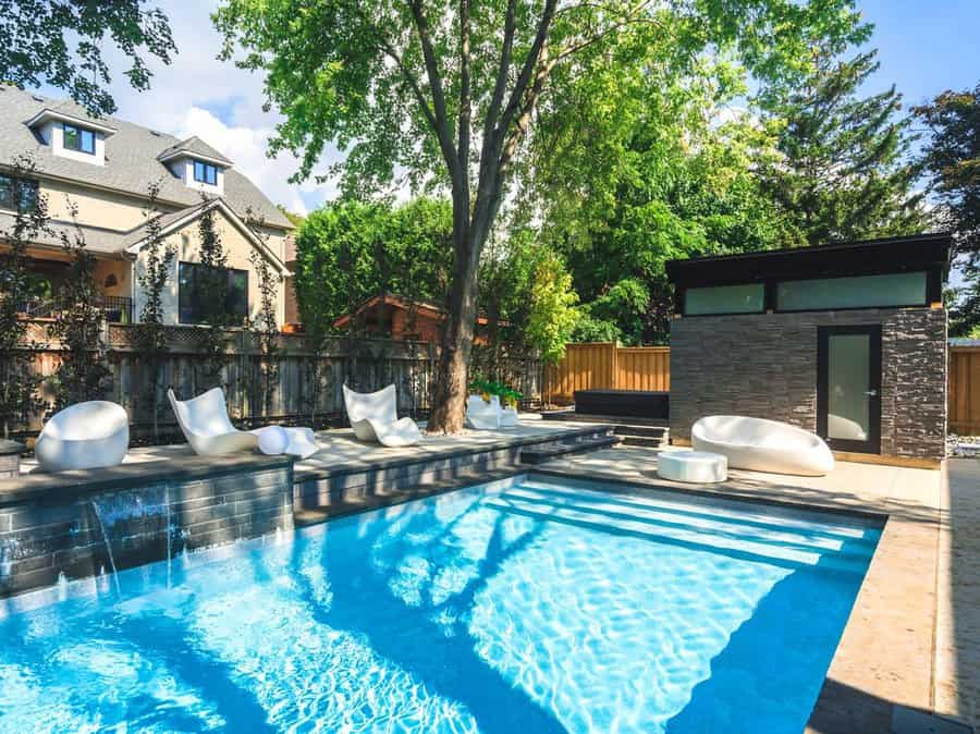 Modern backyard with a pool, stylish white loungers, a wooden deck, and a small contemporary building under a large tree