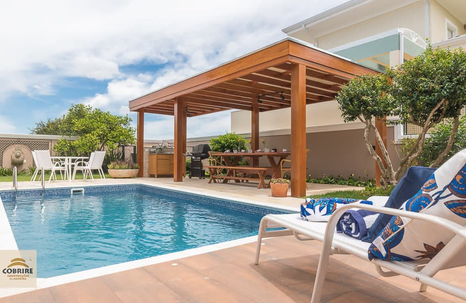 Backyard pool with wooden pergola, surrounded by greenery, lounge chairs, a dining set, and clear skies add to the relaxing atmosphere