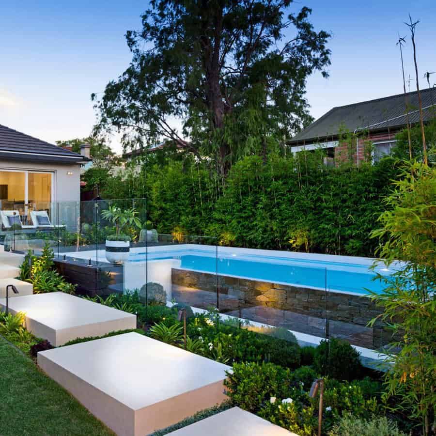 Modern backyard with a glass-enclosed pool, landscaped garden, and a seating area near a house at dusk