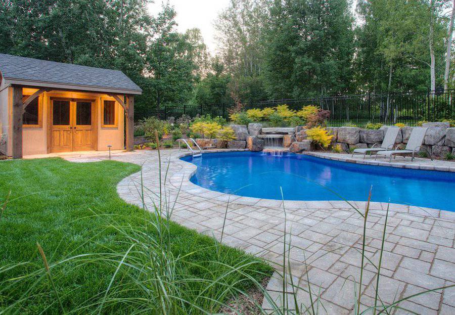 Backyard with a curvy pool, stone patio, wooden pool house, lounge chairs, and surrounding trees and greenery