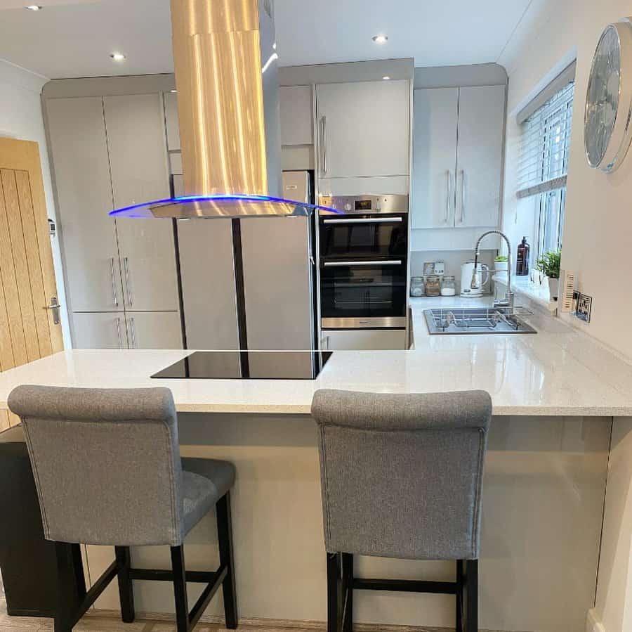 Modern kitchen with a sleek white breakfast bar, gray upholstered barstools, glossy cabinetry, and a striking stainless steel range hood with LED lighting