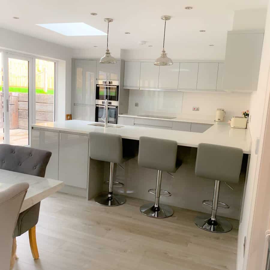 Bright modern kitchen with a sleek white breakfast bar, gray barstools, glossy cabinetry, pendant lighting, and large glass doors for natural light