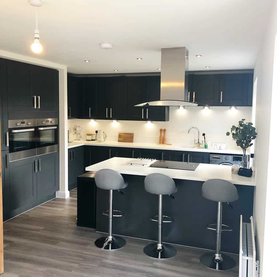 Modern monochrome kitchen with a sleek black breakfast bar, gray barstools, white countertops, stainless steel appliances, and under-cabinet lighting