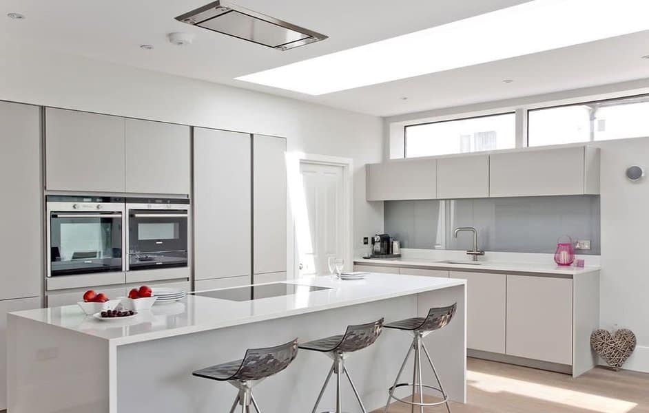 Minimalist white kitchen with a sleek waterfall breakfast bar, glossy cabinetry, transparent barstools, and skylight windows for bright natural lighting