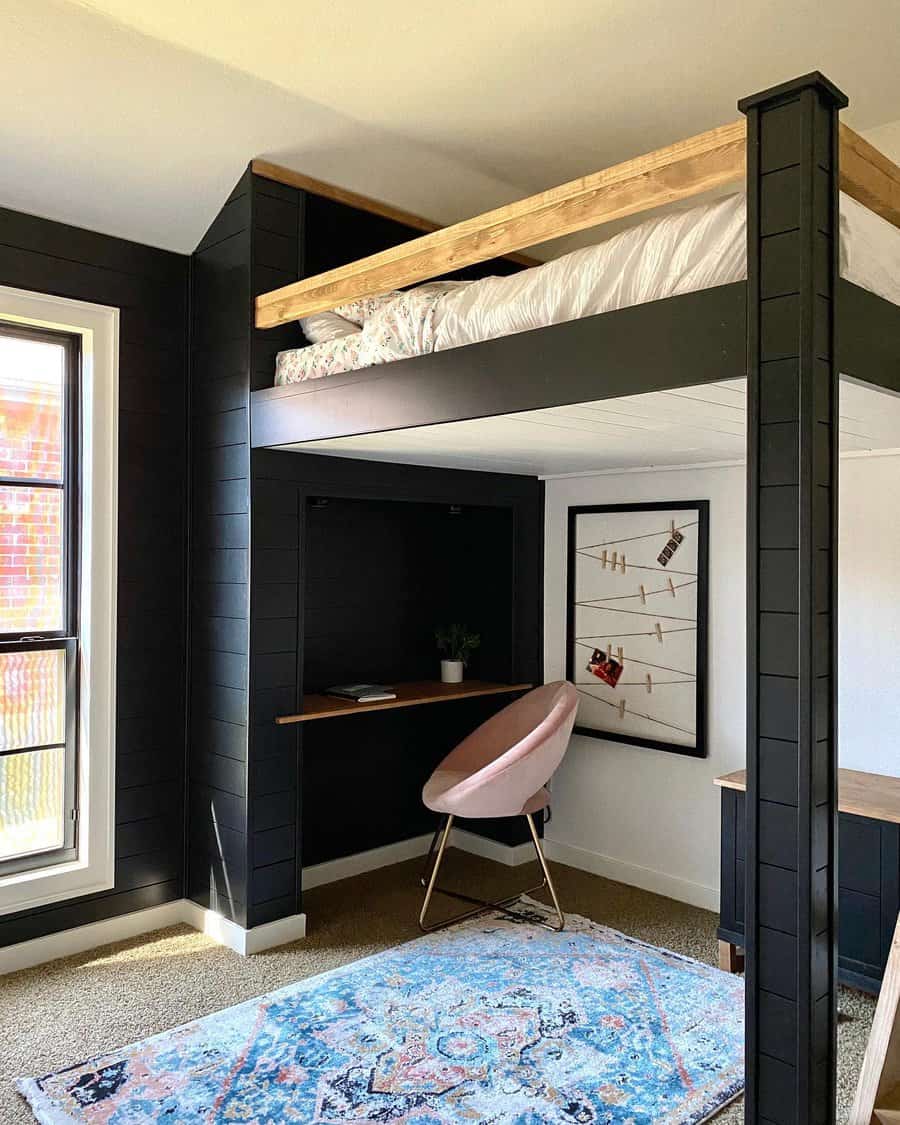 Loft bed with a workspace underneath, featuring a desk, pink chair, and art on the wall; a window with sunlight is on the left