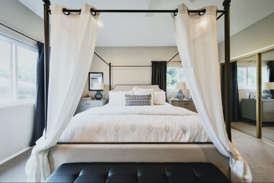 Canopy bed with white curtains, elegant bedding, and bedside lamps in a bright bedroom with large windows and a mirrored closet