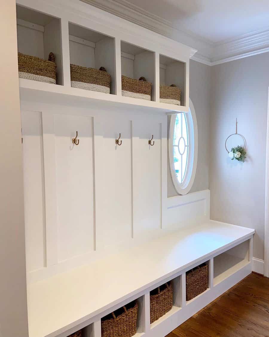 White mudroom with a large bench, wicker baskets on shelves, and brass hooks; oval window and a decorative wall hanging are visible