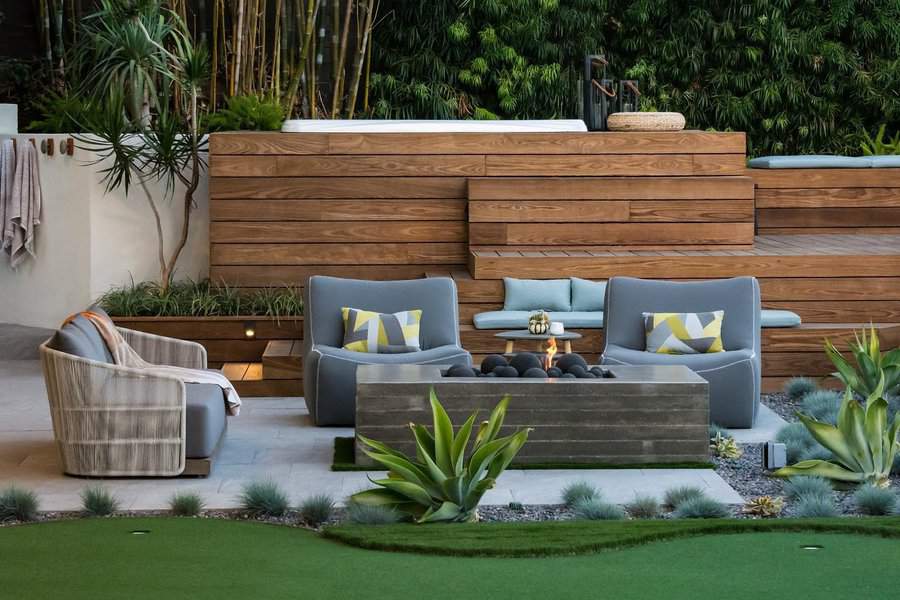 Modern patio with gray chairs, geometric pillows, a sleek fire pit, lush greenery, and terraced wood decking in the background