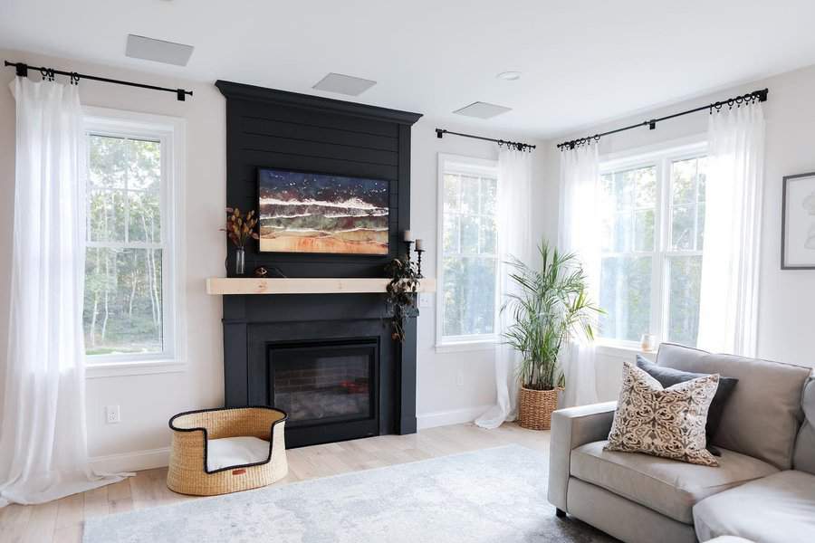 A modern black fireplace with a wooden mantel, featuring a TV above, surrounded by natural light from large windows and minimalist decor