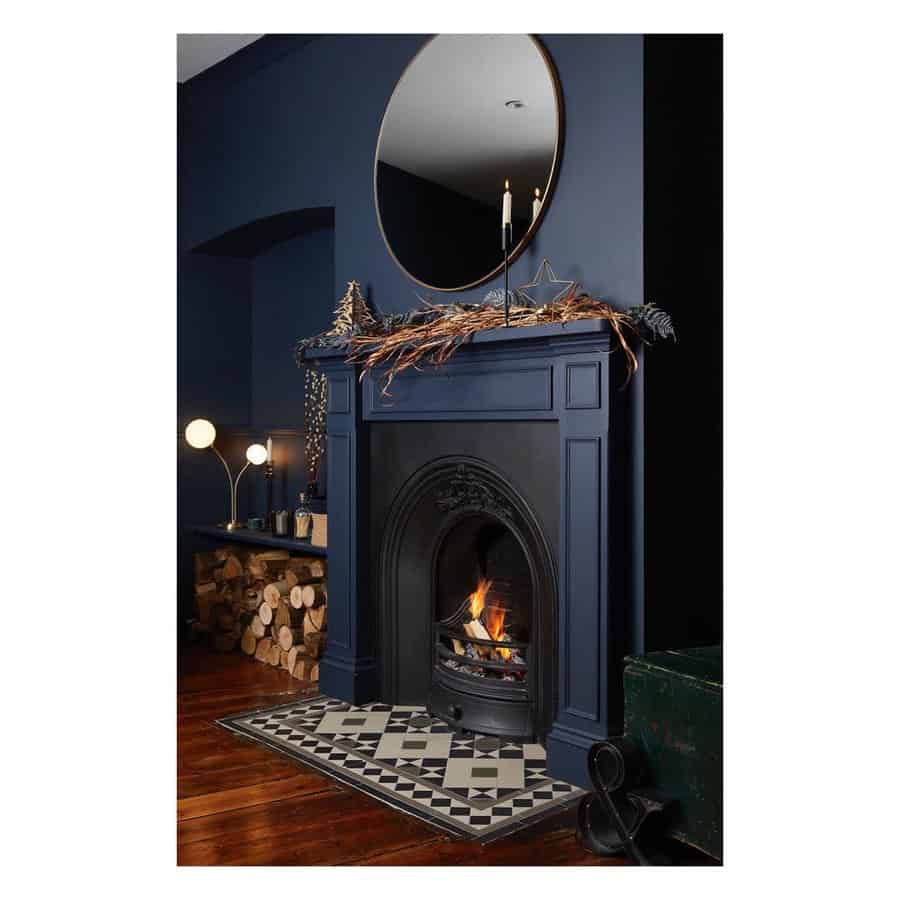 A stylish dark blue fireplace with a geometric patterned rug, a round mirror above, and festive decor, creating a modern and cozy ambiance