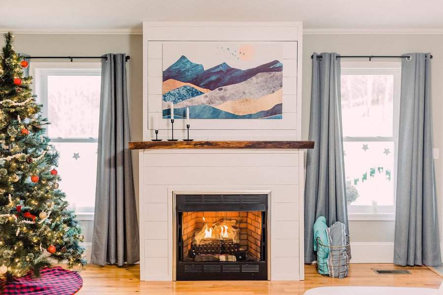 A modern fireplace with a wooden mantel, featuring a colorful mountain painting above, complemented by a Christmas tree and cozy decor for a festive, inviting look