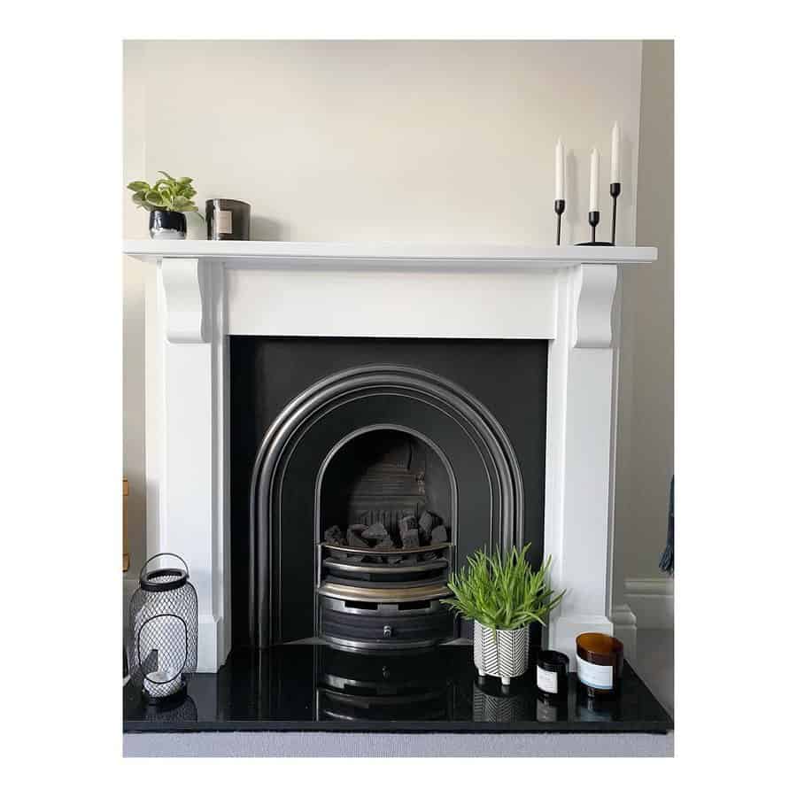 A sleek white fireplace with a black insert, complemented by simple decor like plants, candles, and a lantern, creating a clean and minimalist look