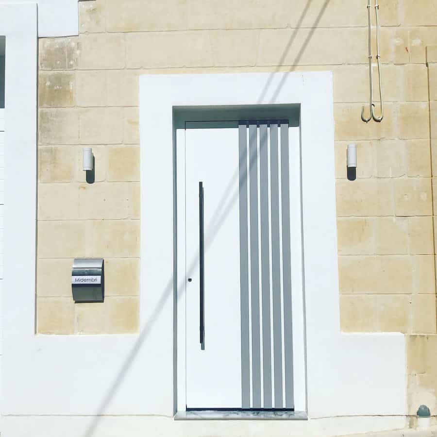 Modern white door with vertical silver stripes on a beige brick wall, flanked by minimalist lights, and a mailbox on the left