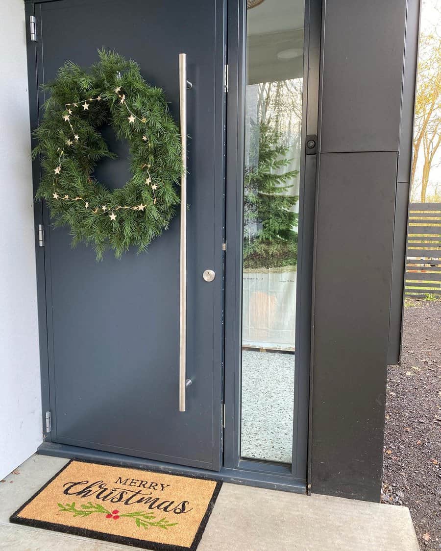 A dark door with a green wreath adorned with small stars. A "Merry Christmas" doormat lies on the ground in front