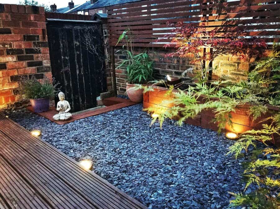 Zen garden with Buddha statue, illuminated plants, slate path, and wooden deck, surrounded by brick and wooden fence