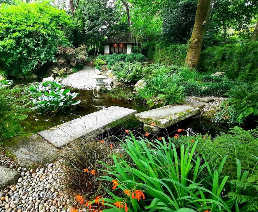 Japanese garden with a small bridge over a pond, stone lantern, lush greenery, orange flowers, and a house in the background