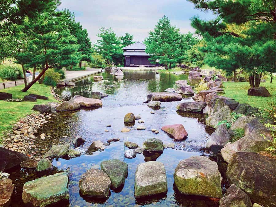 A tranquil Japanese garden with a pond, rocks, lush trees, and a small pavilion in the background under a cloudy sky