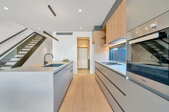 Modern kitchen with sleek cabinetry, built-in oven, stovetop, island with sink, and light wood flooring next to a staircase