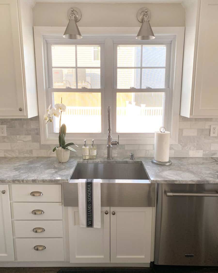 A modern kitchen sink with a stainless steel faucet, orchid plant, soap dispensers, and a towel, under a window with two ceiling lights