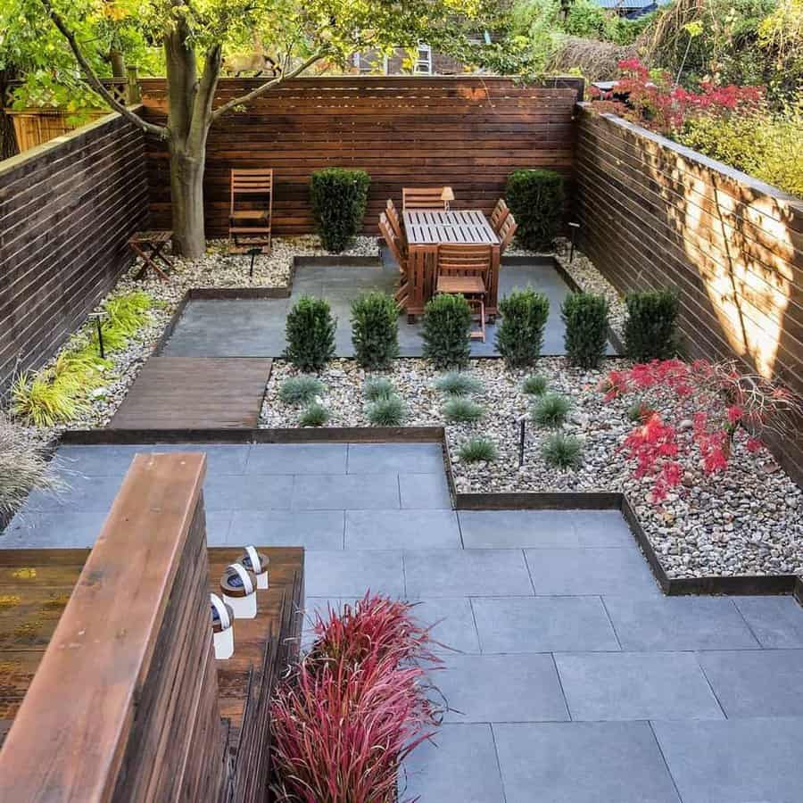 Modern backyard with geometric hardscaping, a wooden dining set, neatly trimmed shrubs, and vibrant plants bordered by wooden fencing