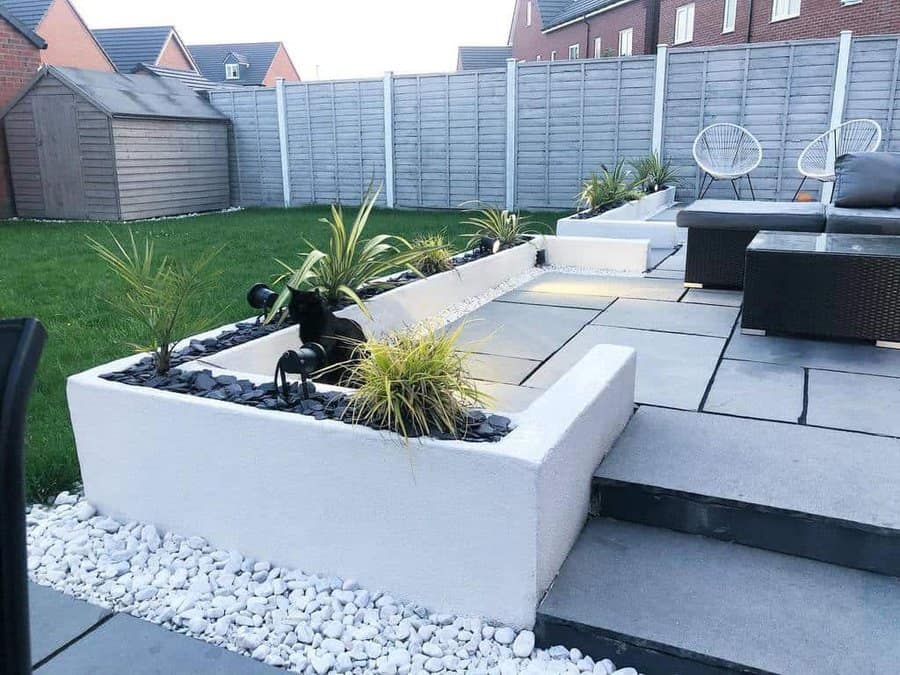 Modern backyard design featuring white raised planters with decorative rocks and greenery, large gray pavers, and outdoor seating against a gray fence backdrop