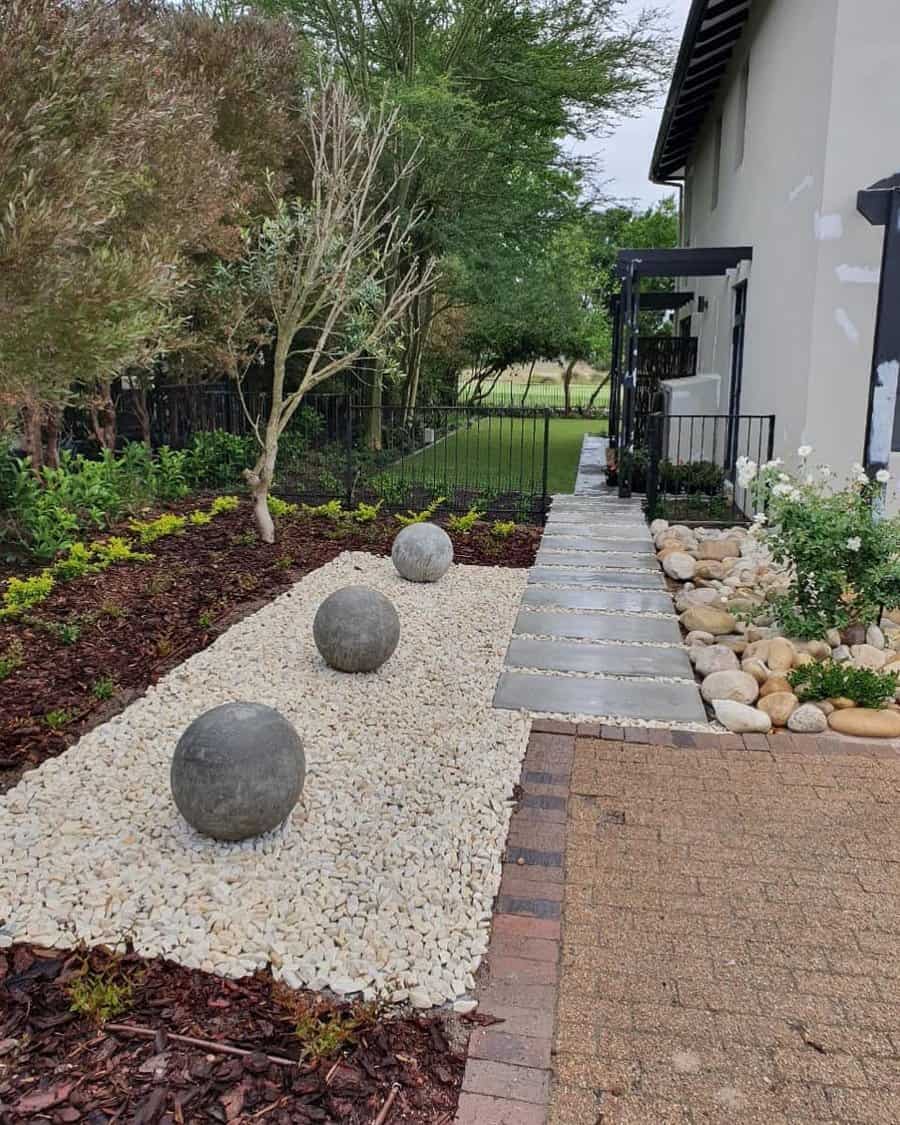 Modern low-maintenance landscaping design featuring smooth concrete spheres on a bed of white pebbles, clean stepping stone pathways, and a blend of mulch, greenery, and natural stone accents for an elegant and structured aesthetic