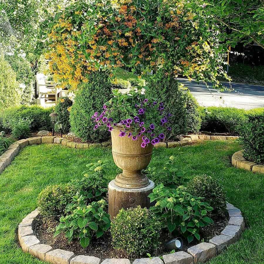 Elegant centerpiece garden design featuring a raised urn filled with vibrant purple petunias, surrounded by lush green boxwood shrubs and seasonal flowers. A natural stone border adds structure, while flowering vines above create a charming canopy effect.