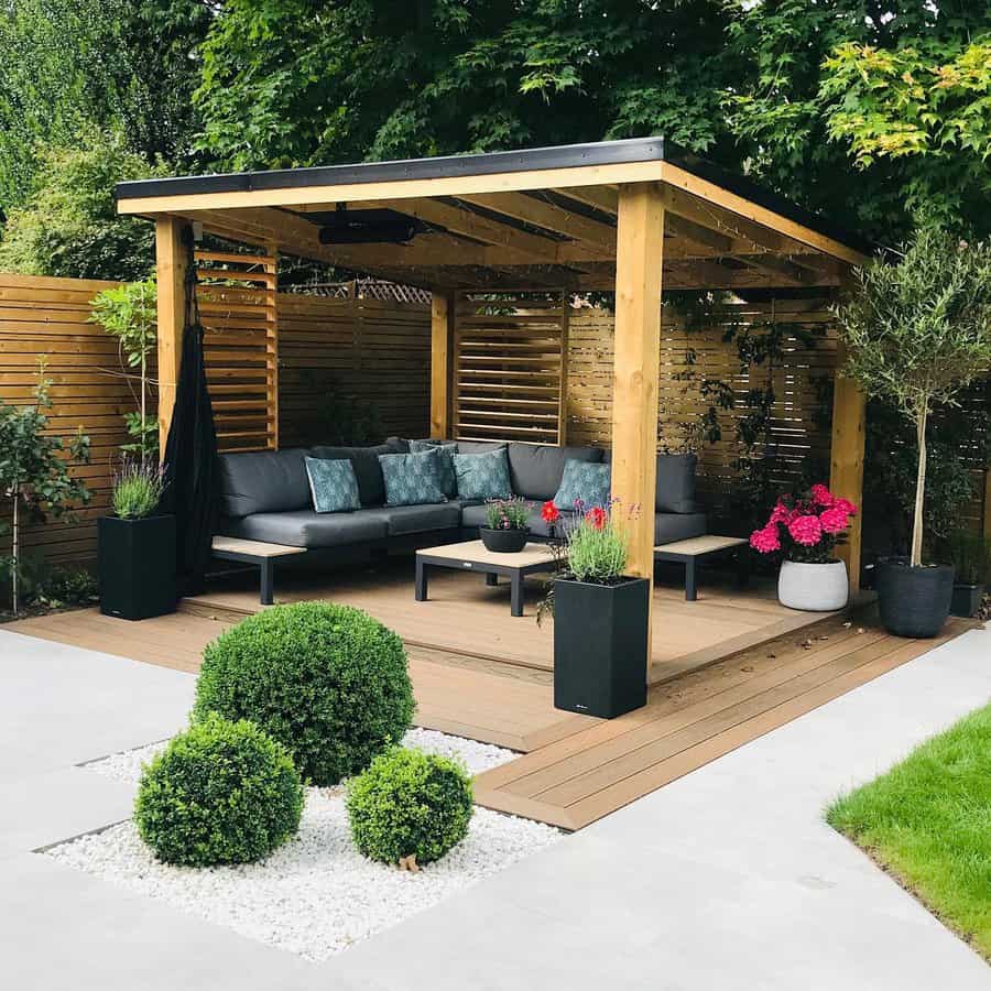 Modern outdoor oasis featuring a sleek wooden pergola with slatted walls, offering a cozy seating area complete with gray cushions and decorative pillows. Surrounding planters with bright blooms and greenery complement the minimalist design, while a trio of manicured boxwoods sits elegantly in a white gravel bed for a touch of sophistication.