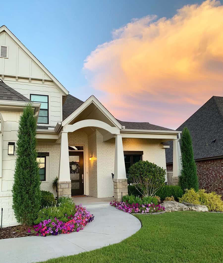 Stunning modern home exterior with a vibrant and low-maintenance landscape. A curved walkway lined with colorful flowers, including bright pink and purple petunias, leads to the welcoming entrance. Towering evergreens and neatly trimmed shrubs add structure, while the sunset sky completes the serene ambiance.