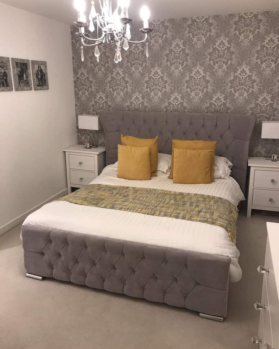 Elegant bedroom with gray tufted bed, golden pillows, and a chandelier; floral wallpaper and white nightstands add a classic touch