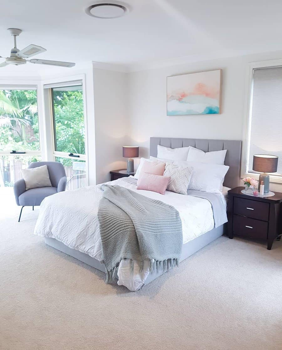 Bright bedroom with a bed, gray headboard, textured blanket, lamps on wooden nightstands, an armchair, and large windows