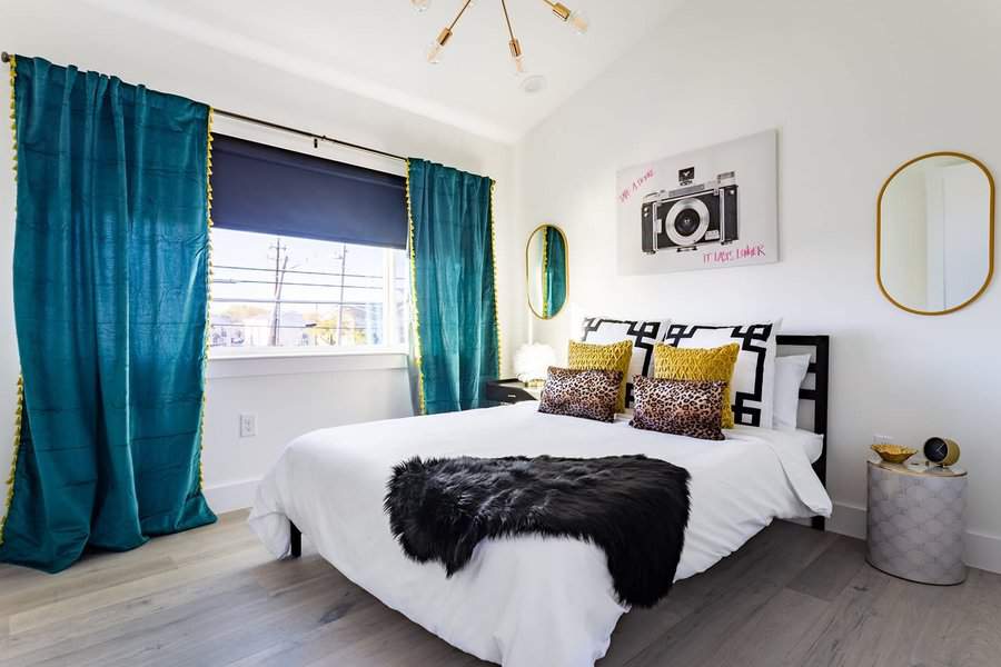 Modern bedroom with a white bed, leopard print and black pillows, teal curtains, camera art on the wall, and a small round side table