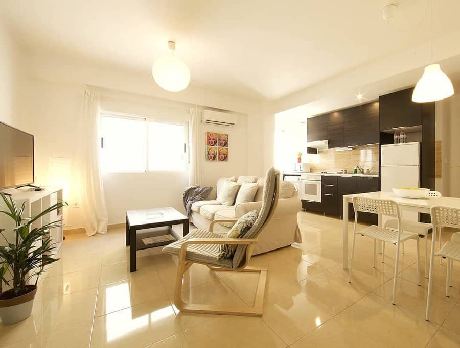 Bright, modern apartment with open living area, dining table, kitchen, TV, sofa, and natural light from a large window