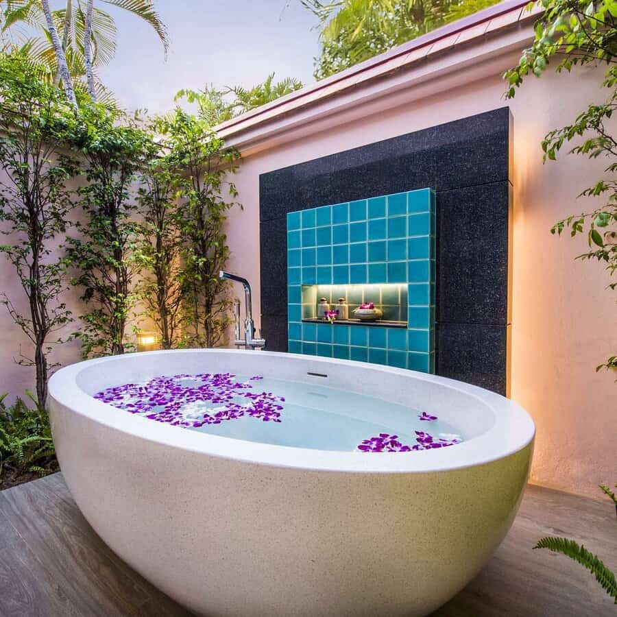 Outdoor bathtub filled with water and purple petals, surrounded by greenery, with a tiled wall feature and small shelf